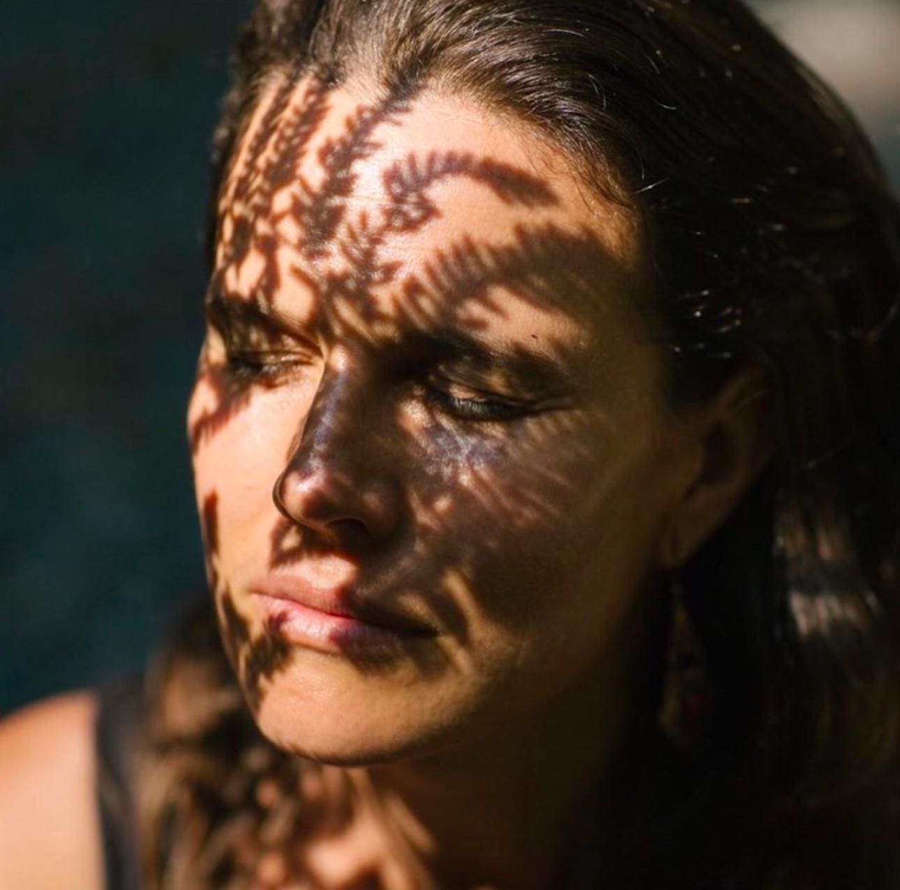 Close-up portrait of a person with intricate fern-like shadows cast across their face and neck, creating a natural pattern. The person has dark hair and is photographed in natural lighting with their eyes gently closed, showing a peaceful expression. The botanical shadows create an artistic interplay of light and dark across their features.